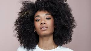 Brown skin woman with long afro headshot against nude background. 